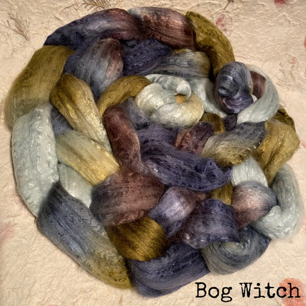 A braided hand dyed IxCHeL cashmerino silk top fluffy, multi-coloured olive gold, slate blue, mauve, cream, and charcoal, displayed on a textured beige fabric background with the text 'Bog Witch' in the bottom right corner.
