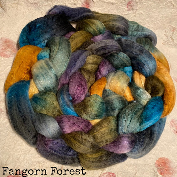 A braided hand dyed IxCHeL cashmerino silk top in a variegated colorway labeled 'Fangorn Forest,' showing blended blue, teal, purple, olive green, and mustard yellow tones on a light textured background.