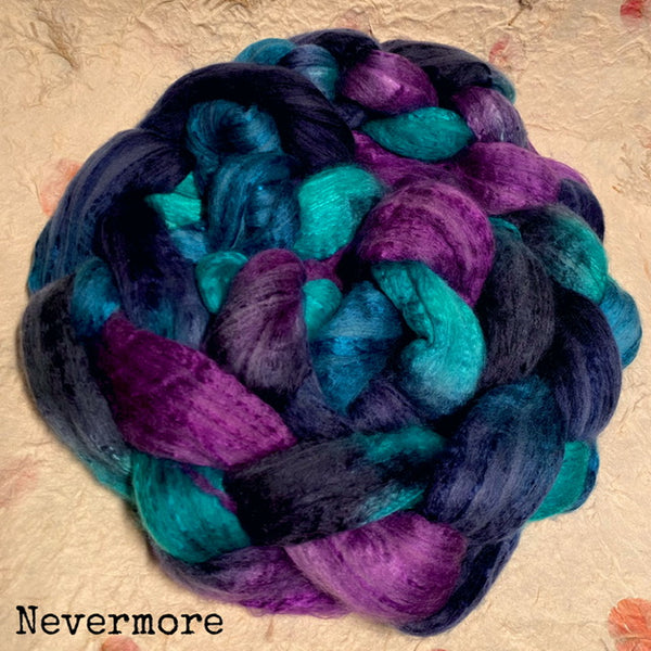A braid of variegated hand dyed IxCHeL cashmerino silk top  in dark navy, teal, and purple, photographed close up on a textured paper background with the word 'Nevermore' printed in the lower left corner.