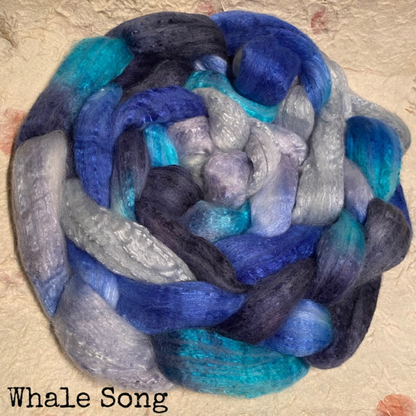 A braid of hand dyed IxCHeL cashmerino silk top in blended blue, teal, blue grey tones , photographed on a textured hand made paper surface. The text 'Whale Song' appears in the lower left corner.