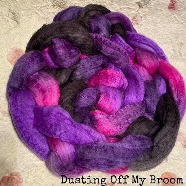 A braided hand dyed IxCHeL cashmerino silk top  in purple, black, and hot pink, resting on a textured cream fabric background with the text 'Dusting Off My Broom' at the bottom.
