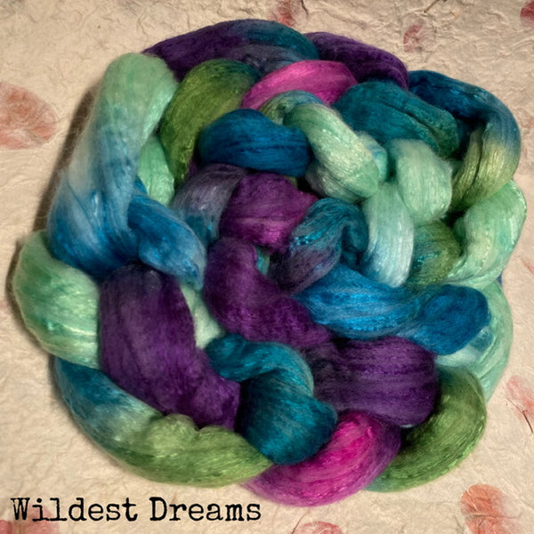 A braided IxCHeL hand dyed  silk top  bundle in shades of purple, teal, mint green, and magenta, displayed on a textured beige fabric background with the brand name 'Wildest Dreams' printed in the bottom-left corner.