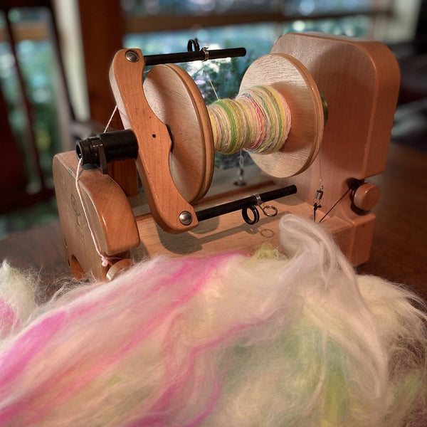 IxCHeL Fibre And Yarns Custom Spinning Service showing a view of a batt of fluff in front to f an Ashford Electric Spinning Wheel