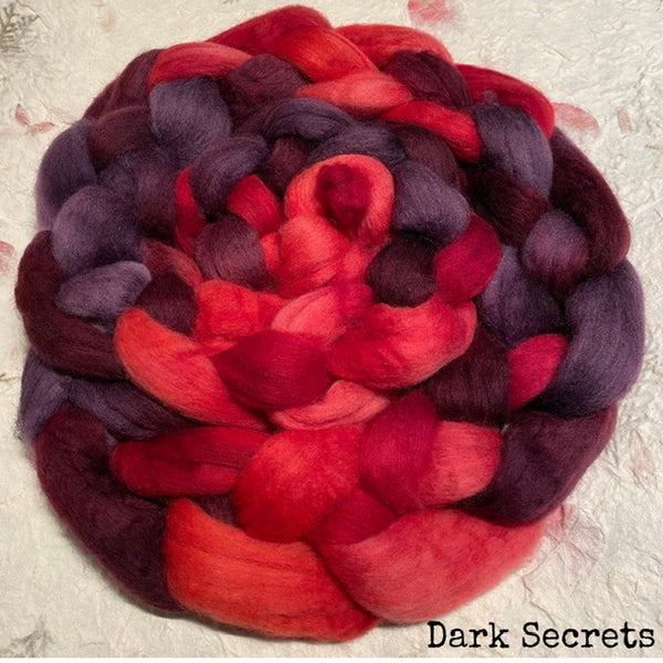 An IxCHeL hand dyed bundle of fluffy Polwarth top roving in a variegated blend of deep red, magenta, and dark purple. The words 'Dark Secrets' appear in the bottom-right corner.