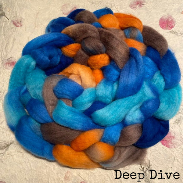 A braid  of IxCHeL hand dyed fluffy Polwarth wool top in a blend of bright blue, turquoise, orange, and taupe-brown, photographed on a textured handmade paper with pink floral marks. The text 'Deep Dive' appears in the lower right corner.