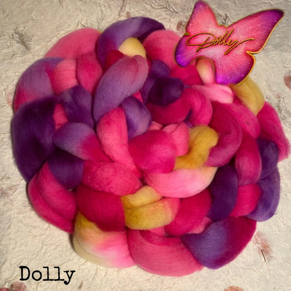 A braided bundle of fluffy handdyed IxCHeL yarns and fibres wool roving top in blended pink, purple, and pale yellow tones, photographed from above on a textured light background. A butterfly-shaped tag with the word Dolly is placed on top, and the word Dolly appears in the lower left corner.