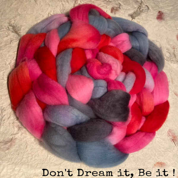 A braided bundle of fluffy wool fibers in blended pink, red, grey, light blue, and dark charcoal tones, photographed on a textured handmade paper background with the text 'Don't Dream it, Be it!' at the bottom.