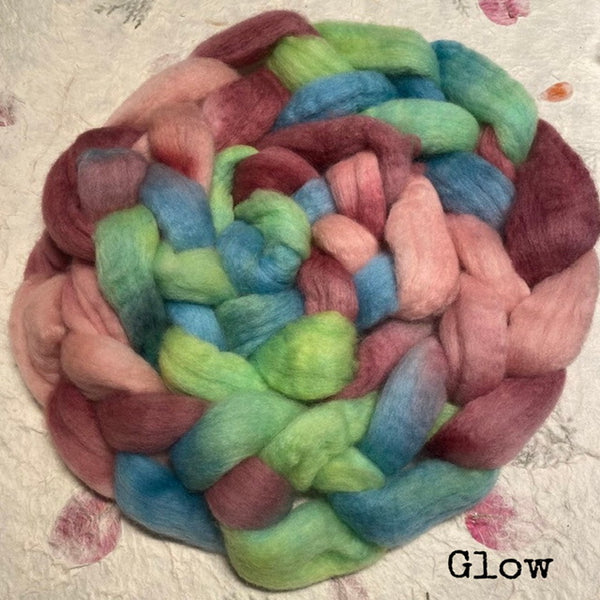 An IxCHeL hand dyed bundle of fluffy Polwarth wool roving with blended shades of pink, green, blue, and burgundy, placed on a handmade paper background with the word 'Glow' in the bottom right corner.