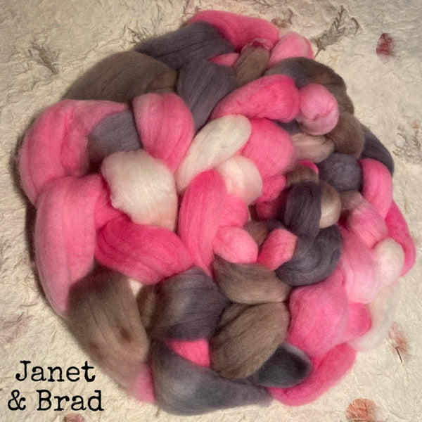 A braided bundle of fluffy wool Polwarth top roving in blended colours, including bright pink, grey, taupe-brown, and off-white, photographed on a textured handmade paper background. The text 'Janet & Brad' appears in the lower-left corner.