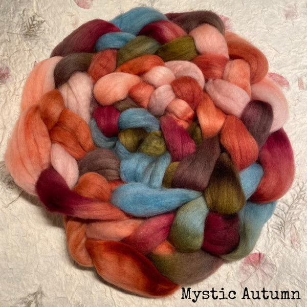 An IxCHeL hand dyed soft Polwarth top roving in a variegated autumn colourway, including coral, rust, pink, blue, olive green, and plum, displayed on a handmade paper with the text 'Mystic Autumn' in the bottom right corner.