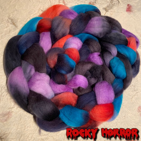 A braided bundle of fluffy Polwarth wool roving tops  in a multicolour blend of black, purple, blue, orange, and red, photographed on a light handmade paper background with a red 'ROCKY HORROR' logo at the bottom.