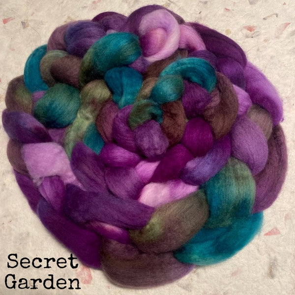 A braid bundle of IxCHeL hand-dyed fluffy Polwarth wool roving top in purple, teal, pink, and olive-brown tones, with the text 'Secret Garden' in the lower left corner.