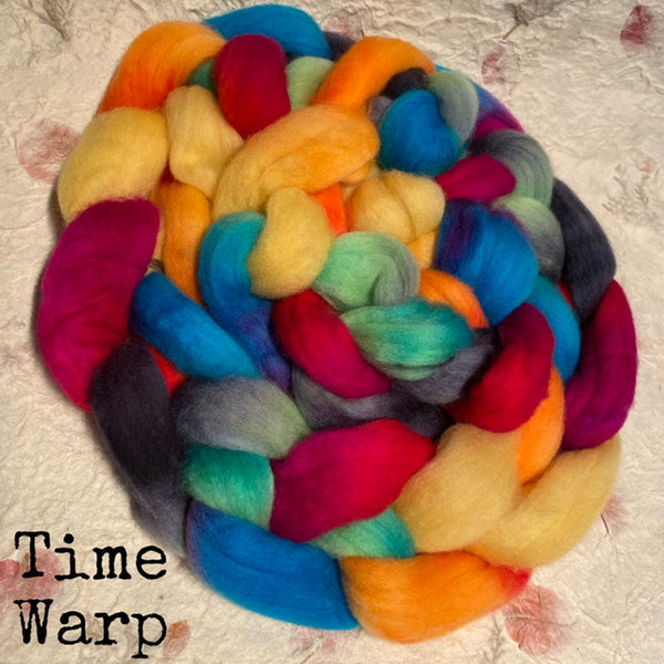 A braided bundle of colourful IxCHeL fibre wool roving top labeled 'Time Warp' in the lower-left corner, photographed on a textured handmade paper background.