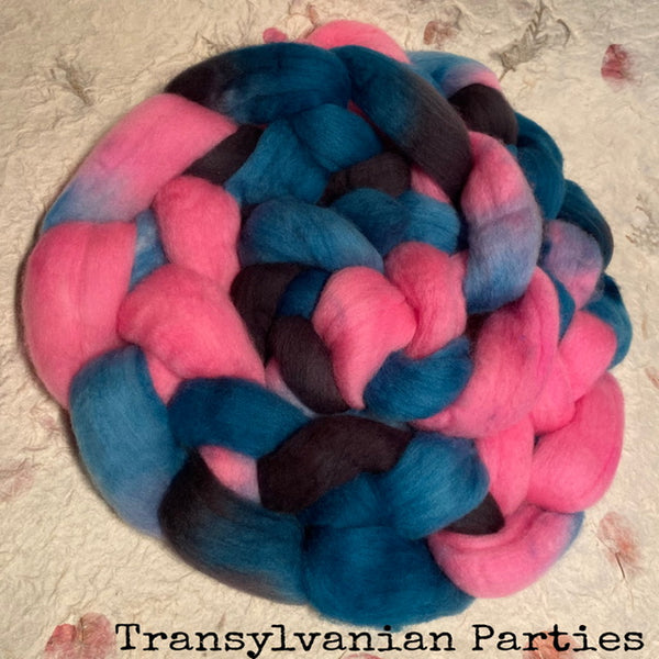 A round braid of fluffy hand dyed IxCHeL yarns and fibres wool roving in bright pink, teal blue, and dark charcoal gray, photographed from above on a textured handmade paper background. The text 'Transylvania Parties' is printed at the bottom.