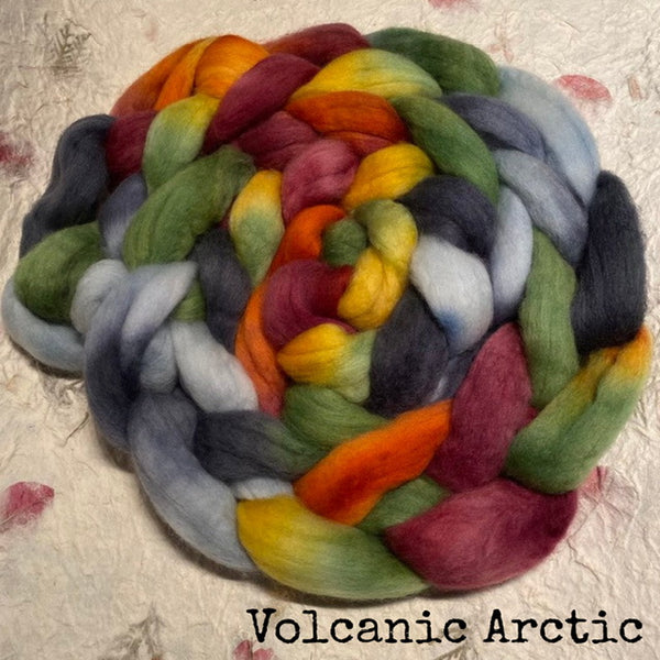 A braid of soft, Polwarth in a blend of IxCHeL handdyed grey, green, yellow, orange, red, and magnolia  tones, placed on a handmade  paper background with the text 'Volcanic Arctic' at the bottom.